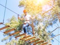 Kid in orange helmet climbing in trees on forest adventure park. Girl walk on rope cables and high suspension bridge in