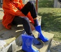 Kid in orange raincoat putting on blue rubby boots sitting on stairs in backyard. Clothing for rainy weather