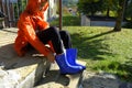 Kid in orange raincoat putting on blue rubby boots sitting on stairs in backyard. Clothing for rainy weather