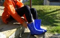 Kid in orange raincoat putting on blue rubby boots sitting on stairs in backyard. Clothing for rainy weather