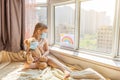 Kid with painted rainbow during Covid-19 quarantine at home. Caucasian Girl with teddy bear toy near window. Stay at Royalty Free Stock Photo