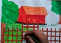 Kid painting a beautiful scenery of hut, trees and green grass holding paint brush in hand