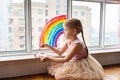 Kid painting rainbow during Covid-19 quarantine at home. Girl near window. Stay at home Social media campaign for coronavirus Royalty Free Stock Photo