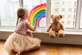 Kid painting rainbow during Covid-19 quarantine at home. Girl near window. Stay at home Social media campaign for coronavirus Royalty Free Stock Photo