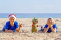 Kid in red Santa hats and pineapple as Christmas tree lying at sea beach during Christmas vacation