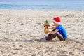 Kid in red Santa hats and pineapple as Christmas tree sitting at sea beach during Christmas vacation