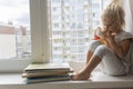 Kid sits near window, holding bear toy and looking at books at windowsill. Royalty Free Stock Photo