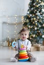 The kid, sitting on the floor near a smart Christmas tree, collects a multicolored pyramid. Royalty Free Stock Photo