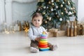 The kid, sitting on the floor near a smart Christmas tree, collects a multicolored pyramid. Royalty Free Stock Photo