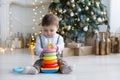 The kid, sitting on the floor near a smart Christmas tree, collects a multicolored pyramid. Royalty Free Stock Photo