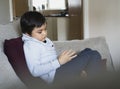 Kid sitting on sofa watching cartoon on tablet, Sad child sitting alone looking down deep in thought, Boy relaxing at home playing Royalty Free Stock Photo