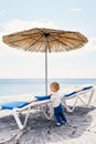 Kid stands leaning on a sun lounger under a thatched beach umbrella and looks at the sea Royalty Free Stock Photo