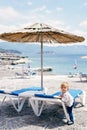 Kid stands near a sun lounger under a straw umbrella on the beach near the water Royalty Free Stock Photo