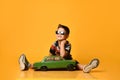 Kid in sunglasses, camouflage t-shirt, sneakers. Sitting on floor, holding green model of retro car, posing on orange background