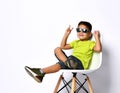 Kid in sunglasses, yellow t-shirt, denim shorts, khaki sneakers. Smiling showing victory sign, sitting on chair.  on white Royalty Free Stock Photo