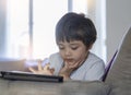 Kid touching finger on tablet screen, Child boy lying on sofa relaxing at home watching cartoons or playing games on digital pad, Royalty Free Stock Photo