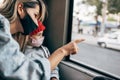 The kid wears a protective face mask in red winter hat looking into the window while going to school by bus with her mother. Royalty Free Stock Photo