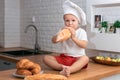 Kid in a white chef`s hat in the kitchen eating a baguette, little mother`s helper, boy cook, concept of family weekend, delicious