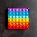 Kids anti stress sensory pop it or simple dimple fidget push toy on a black table or floor background. square popit rainbow hue br