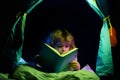 Kids in kids tent reading a book in the dark home. Child boy reading a book lying on the bed. Dreaming child read Royalty Free Stock Photo