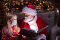 Kids and Santa in medical masks. Girl and Santa Claus reading Christmas book sitting near Christmas tree at home. Happy Royalty Free Stock Photo