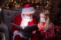 Kids and Santa in medical masks. Girl and Santa Claus reading Christmas book sitting near Christmas tree at home. Happy Royalty Free Stock Photo