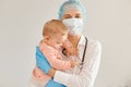 Kind hearted woman doctor pediatrician wearing medical cap, surgical mask, gown and rubber gloves, standing with toddler girl in Royalty Free Stock Photo