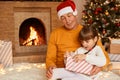 Kind senior man wearing orange sweater and santa claus hat posing with little girl opening his Christmas present with surprised