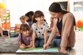 Kindergarten teacher reading book with cute girls while other children playing together Royalty Free Stock Photo