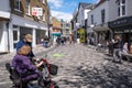 Disabled Woman In A Wheelchair With Her Carer In A Sunny High Street With Shoppers
