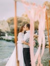 The kissing newlyweds behind the wedding arch decorated with pink ribbons at the background of the sea.