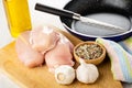 Knife in frying pan, vegetable oil, pieces of raw chicken breast, garlic, bowl with condiment on cutting board on table Royalty Free Stock Photo