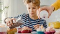 In the Kitchen: Portrait of the Smart Little Boy Sprinkling Funfetti on Creamy Cupcakes Frosting. Royalty Free Stock Photo