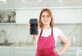 in kitchen,smiling red-haired girl in polka-dot apron shows blank smartphone screen close-up