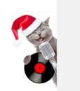 Kitten with retro microphone with vinyl record in red santa hat peeking from behind empty board. isolated on white background Royalty Free Stock Photo