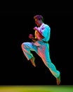 Knee kick. Young man in white kimono practicing karate punches, kicks over black studio background in neon lights