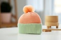Knitted baby hat in pastel orange green colours with pompon on white table
