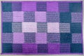 Knitted factory fabric floor mat of alternating squares of purple, violet, blue, lilac and pink laid out on the concrete floor