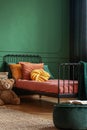 Knot yellow pillow on single metal bed with dark orange bedding, copy space on empty dark green wall