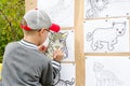 Boy in a baseball cap paints a picture of a leopard that is mounted on a wooden stand