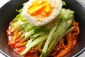 Korean cold noodles with spicy sauce, topped with cucumber, boiled egg and crushed sesame seeds in a black bowl.