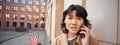 Korean girl with concerned face talks on mobile phone and shrugs, frowns and looks concerned, being lost in town, stands