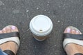 A kraft disposable takeaway paper cup with white cup on asphalt road between woman legs feet wearing leather sandals, summertime Royalty Free Stock Photo