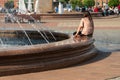 A young woman sits alone at the edge of a fountain in the Railway Station Square filled with people on a hot summer day