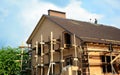 A brick house with scaffolding under construction. Roofers are installing asphalt shingles on the