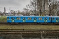 Side view of a blue subway train with yellow stripes, outdoors on a cloudy autumn day