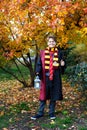 Cute boy in glasses stands in autumn park with gold leaves, holds book in his hands, wears in black robe