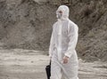 Lab technician in a mask and chemical protective suit, walks on dry ground with a tool box through toxic smoke