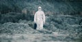 Lab technician in a mask and chemical protective suit, walks on dry ground with a tool box through toxic smoke