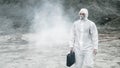 Lab technician in a mask and chemical protective suit, walks on dry ground with a tool box through toxic smoke